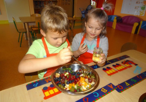 Dwoje dzieci siedzi przy stole. Chłopiec miesza łyżką miód z owocami, dziewczynka przygląda się z zaciekawieniem.